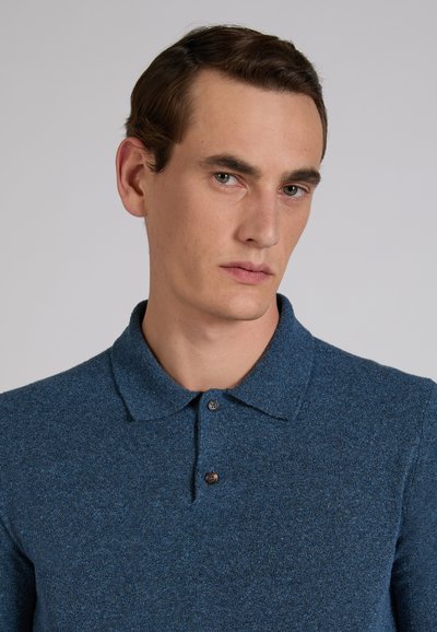 Młody mężczyzna z krótkimi brązowymi włosami, ubrany w niebieską koszulkę polo z guziczkami, patrzący lekko na bok na jednolitym jasnym tle.