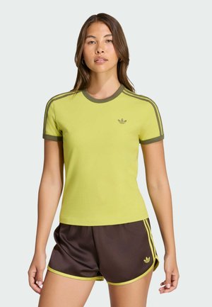 Mujer con camiseta lime green de Adidas con ribete marrón y pantalones cortos marrones de Adidas con ribete lime green, de pie contra un fondo liso.