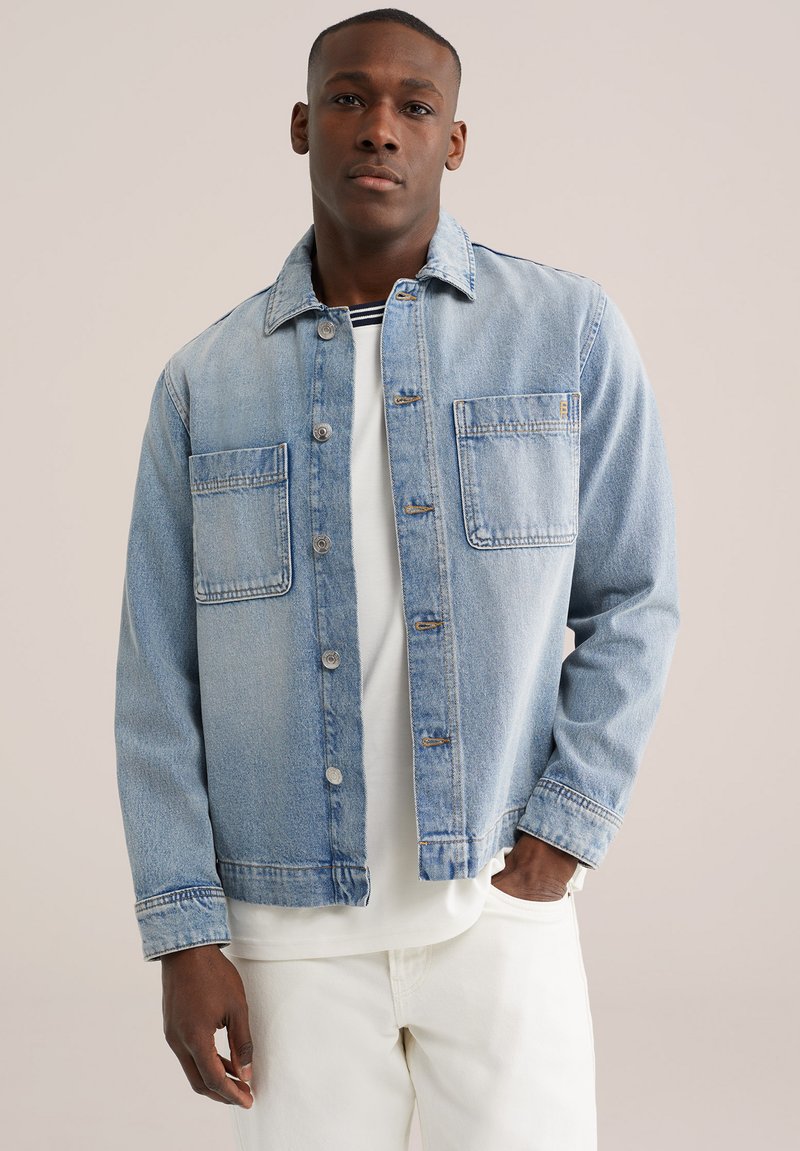 Bărbat îmbrăcat într-o jachetă de denim albastru deschis peste un tricou alb și pantaloni, stând cu o mână în buzunar, pe un fundal simplu.