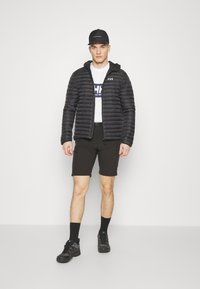 Černá nadýchaná bunda s kapucí, s vertikálním prošíváním. Spárována s bílým tričkem a černými šortkami, spolu s černými teniskami.