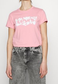 Světle růžové bavlněné tričko s krátkými rukávy, které má na hrudi bílé logo Levi's, v kombinaci se šedými džíny.