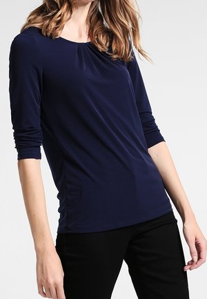 Femme portant un haut bleu marine à manches longues avec un détail de col froncé et un pantalon noir, posant devant un fond clair uni.