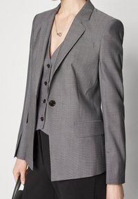 Le blazer gris présente une coupe ajustée, des revers crantés et deux poches avant. Il a une texture subtile et des boutons sombres en guise de fermeture.