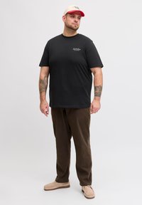 Άνδρας με γενειάδα φοράει μαύρο μπλουζάκι, καφέ παντελόνι, μπεζ παπούτσια, μπεζ και κόκκινο καπέλο, στέκεται μπροστά σε απλό φωτεινό φόντο, με τατουάζ στα αντιβράχια.