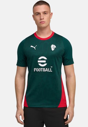 Giovane uomo che indossa una maglia da calcio verde scuro e rossa con logo Puma ed emblema bianco di calcio, in piedi davanti a uno sfondo grigio uniforme.
