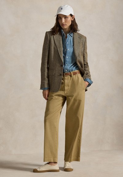 Femme portant une casquette de baseball blanche, un blazer en tweed marron, une chemise en jean bleu, un pantalon beige et des baskets beige, debout devant un fond neutre.
