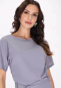Grijze blouse met korte mouwen, ronde halslijn en geplooide taille-detail, gecombineerd met zilveren oorbellen met kralen. Soepele textuur.