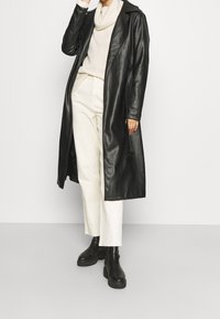 Schwarzer Trenchcoat aus Leder, langes Design, mit Reverskragen. Getragen über einem cremefarbenen Pullover und beigen Hosen. Schwarze Stiefel mit klobiger Sohle vervollständigen den Look.