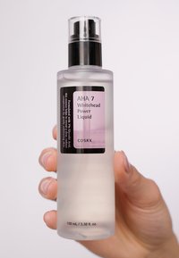 Bouteille transparente et givrée de AHA 7 Whitehead Power Liquid. Étiquette noir et rose avec du texte. Pompe claire en haut ; tenue par une main. Taille de 100 mL.
