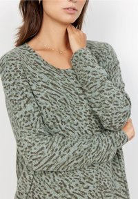 Grüner Pullover mit langen Ärmeln, der mit dunklen, braunen, abstrakten Flecken gemustert ist; weiche Textur; mit rundem Halsausschnitt und gerippten Bündchen.