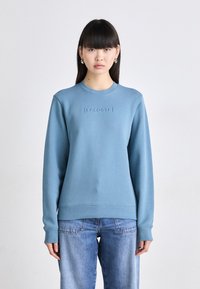 Sudadera azul claro con cuello redondo, puños y dobladillo acanalados, y un logotipo en relieve de "LACOSTE" en la parte delantera. Tejido de mezcla de algodón.