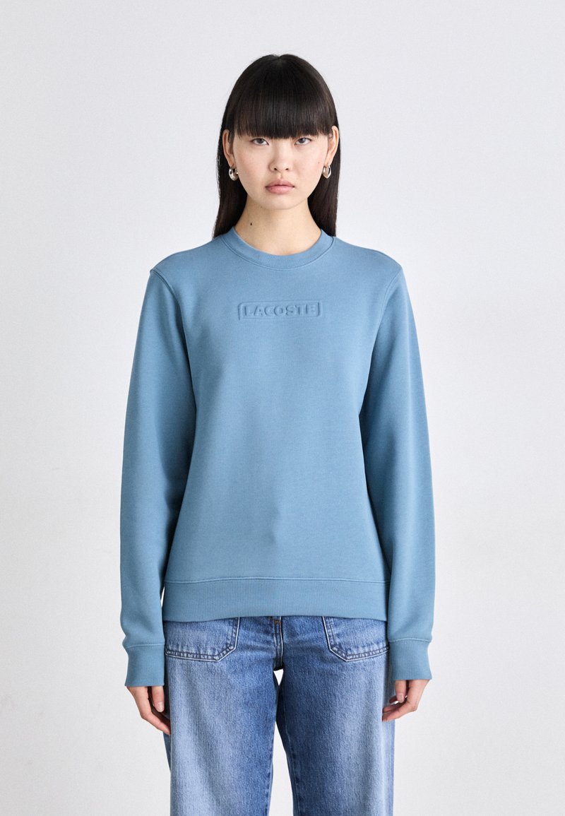 Sudadera azul claro con cuello redondo, puños y dobladillo acanalados, y un logotipo en relieve de "LACOSTE" en la parte delantera. Tejido de mezcla de algodón.
