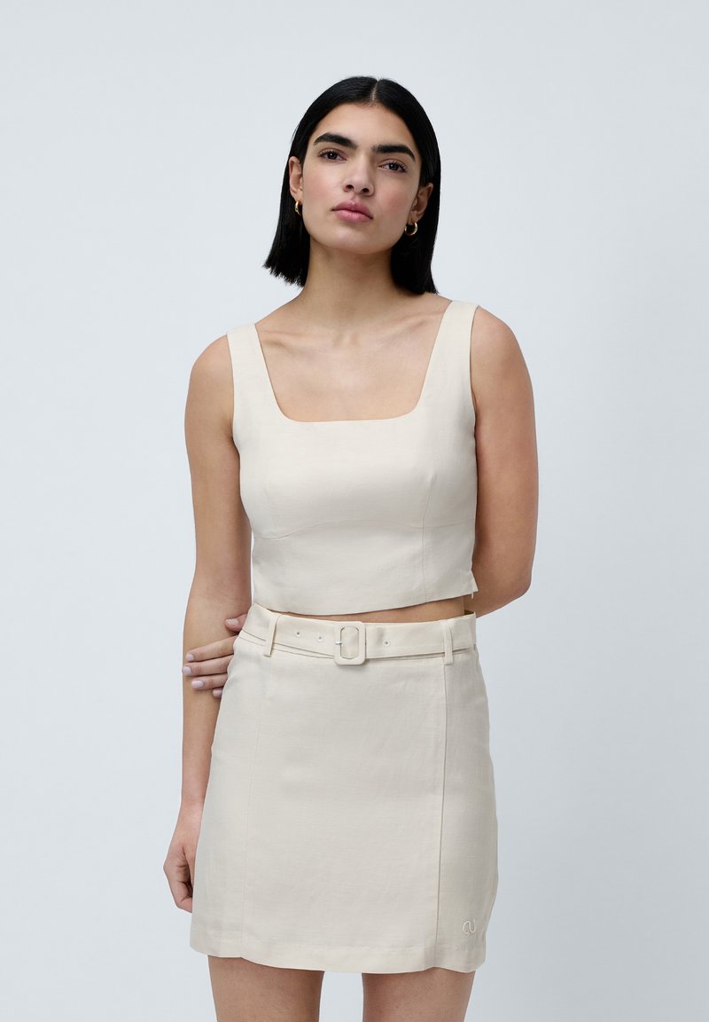 Beige crop top med firkantet halsudskæring, parret med en matchende højtaljet mini-nederdel med bælte og syninger. Glat tekstur.