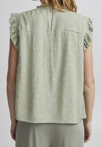 Blusa verde clara con mangas con volantes, tela texturizada y puntos bordados dispersos. Cierre de botón en la parte posterior. Corte suave y relajado.