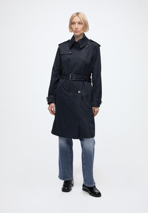 Giovane donna che indossa un trench nero con cintura sopra jeans blu e scarpe nere, in piedi contro uno sfondo bianco uniforme.