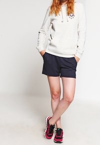 Personne portant un sweat à capuche gris clair avec un motif de cœur, un short bleu marine et des baskets rose vif et noires, debout les jambes croisées.