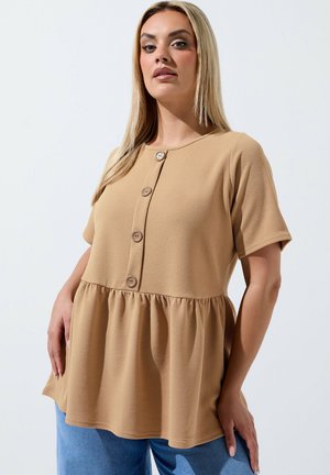 Lyshåret kvinde iført en tan kortærmet knappet peplum top og blå jeans, stående mod en ensartet lys baggrund.