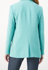 Dlouhý, přizpůsobený blazer z jemné tyrkysové tkaniny. Mezi jeho prvky patří rozparek vzadu, výstřih s klopami a jedno logo poblíž výstřihu.