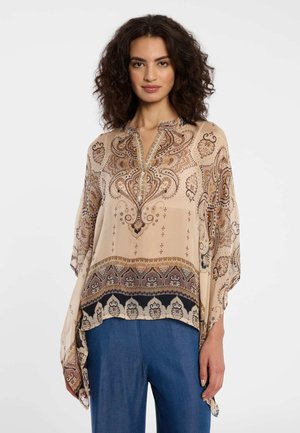 Frau mit lockigem Haar, die eine beigefarbene Bluse mit Paisley-Muster und blaue Hosen trägt, stehend vor einem schlichten hellen Hintergrund.