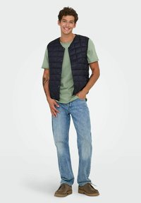 Gilet matelassé bleu marine sur un t-shirt vert clair, associé à un jean bleu et des chaussures marron. Détails notables : boutons sur le gilet.