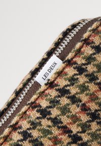 Puzdro so zipsom z vlnenej zmesi s vzorom houndstooth, obsahujúce odtiene béžovej, zelenej, čiernej a červenej; značková etiketa viditeľná pri zips.