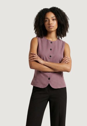 Donna con capelli ricci che indossa una canottiera color malva con bottoni e pantaloni neri, in piedi con le braccia incrociate davanti a uno sfondo uniforme.