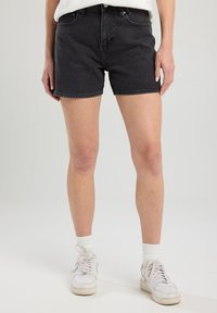 Shorts en denim noir avec une coupe classique, dotés d'une taille haute, d'un design à cinq poches et de coutures visibles. Portés avec des baskets blanches et des chaussettes.