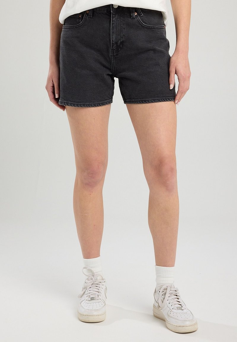Shorts en denim noir avec une coupe classique, dotés d'une taille haute, d'un design à cinq poches et de coutures visibles. Portés avec des baskets blanches et des chaussettes.