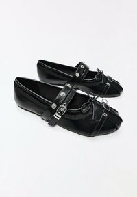 Paire de chaussures plates noires avec brides à boucle et petits nœuds décoratifs sur fond blanc.
