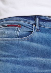 Närbild av en ficka på blå denimjeans med vita, röda och marinblå randiga detaljer, bärs med en vit skjorta instoppad.