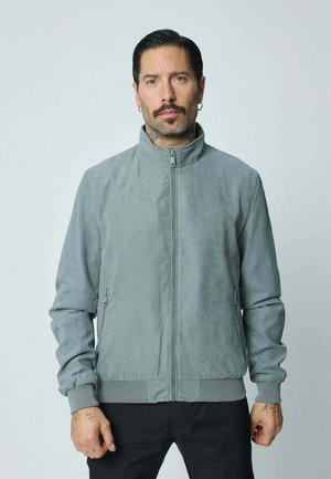 Homme avec moustache et boucles d'oreilles portant une veste zippée gris clair et un pantalon noir, debout devant un fond clair uni.