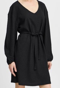 Robe noire texturée avec manches longues et décolleté en V. Elle possède une ceinture à la taille. Le matériau présente une surface froissée pour un détail supplémentaire.