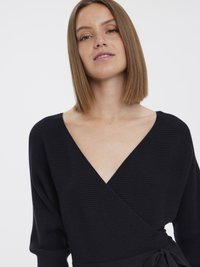 Mulher com cabelo liso, até aos ombros, castanho-claro, vestindo um suéter preto canelado com um decote em V profundo.