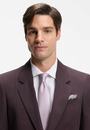 Junger Mann mit braunen Haaren, bekleidet mit einem dunkelvioletten Anzug, weißem Hemd, hellvioletter gemusterter Krawatte und grauem gemustertem Einstecktuch vor weißem Hintergrund.