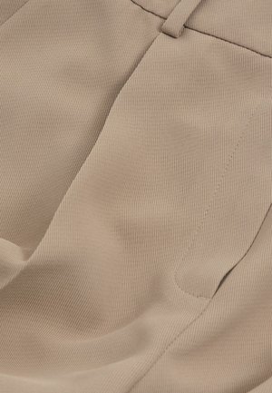 Primer plano de pantalones de tela beige que muestran la cinturilla, el pasador del cinturón y un bolsillo lateral con detalles de costura visibles.