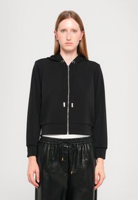 Korte zwarte ritshoodie gemaakt van zachte stof, met een capuchon, zilveren rits en koord, gecombineerd met losse zwarte broek.