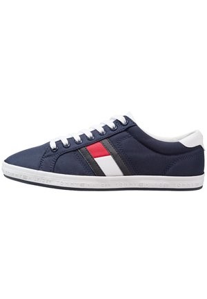 Basket basse bleu marine avec semelle et lacets blancs, présentant un motif de drapeau rouge, blanc et noir sur le côté et texte de la marque sur la semelle.