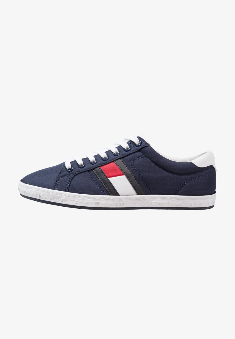 Basket basse bleu marine avec semelle et lacets blancs, présentant un motif de drapeau rouge, blanc et noir sur le côté et texte de la marque sur la semelle.