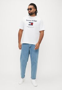 White cotton t-shirt with "Tommy Hilfiger" logo and flag graphic. Paired with light blue denim jeans and white sneakers. Dark sunglasses worn.