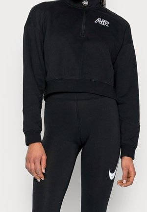 Vrouw draagt zwarte Nike sweatpants met wit swoosh-logo en zwart cropped top met lange mouwen met "AIR" logo en halve rits aan de voorkant.