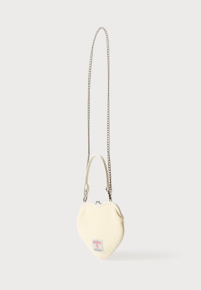 Bolso con forma de corazón en tela color crema con superficie texturizada. Cadena y asa superior en tono plateado, con una pequeña etiqueta con logo en el frente.