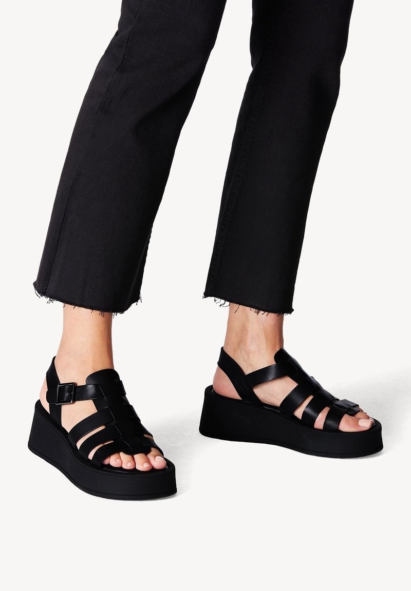 Pieds portant des sandales à plateforme noires avec larges brides et boucles, associées à un pantalon noir raccourci aux ourlets effilochés.
