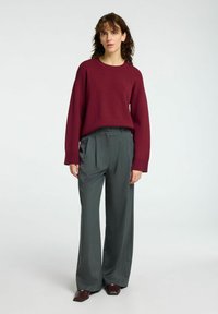 Burgunderfarbener Strickpullover kombiniert mit grauen, gefalteten Hosen. Der Pullover hat eine lockere Passform; die Hose hat ein Streifenmuster. Dunkelbraune Schuhe.