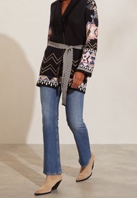 Svart stickad cardigan med färgglada blommönster, med v-ringning, midjebälte och fransade ärmar; ihop med blå jeans och beige anklar stövlar.