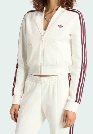 Vrouw draagt een witte zip-up trainingsjas en broek met bordeauxrode strepen langs de mouwen en broekspijpen, staand met ontspannen armen.