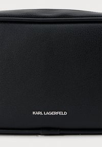 Melna ādas soma ar teksturētu virsmu, kurai ir sudraba "KARL LAGERFELD" logo. Vienkāršs, taisnstūra dizains ar minimālu aparatūru.