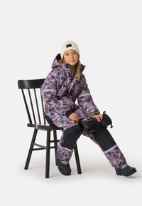 Gewatteerd sneeuwpak in paars met kleurrijke patronen, zilveren accenten en zwarte details. Gecombineerd met een witte gebreide muts en zwarte laarzen.