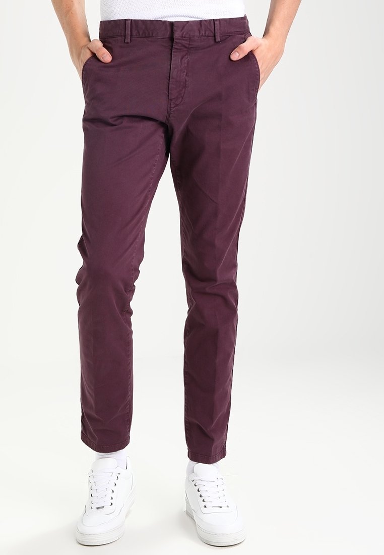 Homme portant un chino violet foncé avec les mains dans les poches et des baskets blanches sur un fond blanc uni.