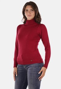 Maglione a collo alto rosso realizzato in tessuto morbido, con dettagli a coste sul colletto e sull'orlo, indossato con jeans blu scuro.