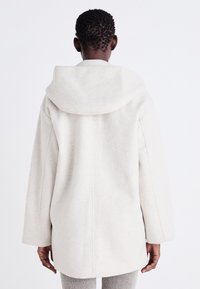 Manteau en laine beige clair avec une capuche spacieuse, des manches larges et une coupe droite avec des coutures minimales et une finition douce et texturée.